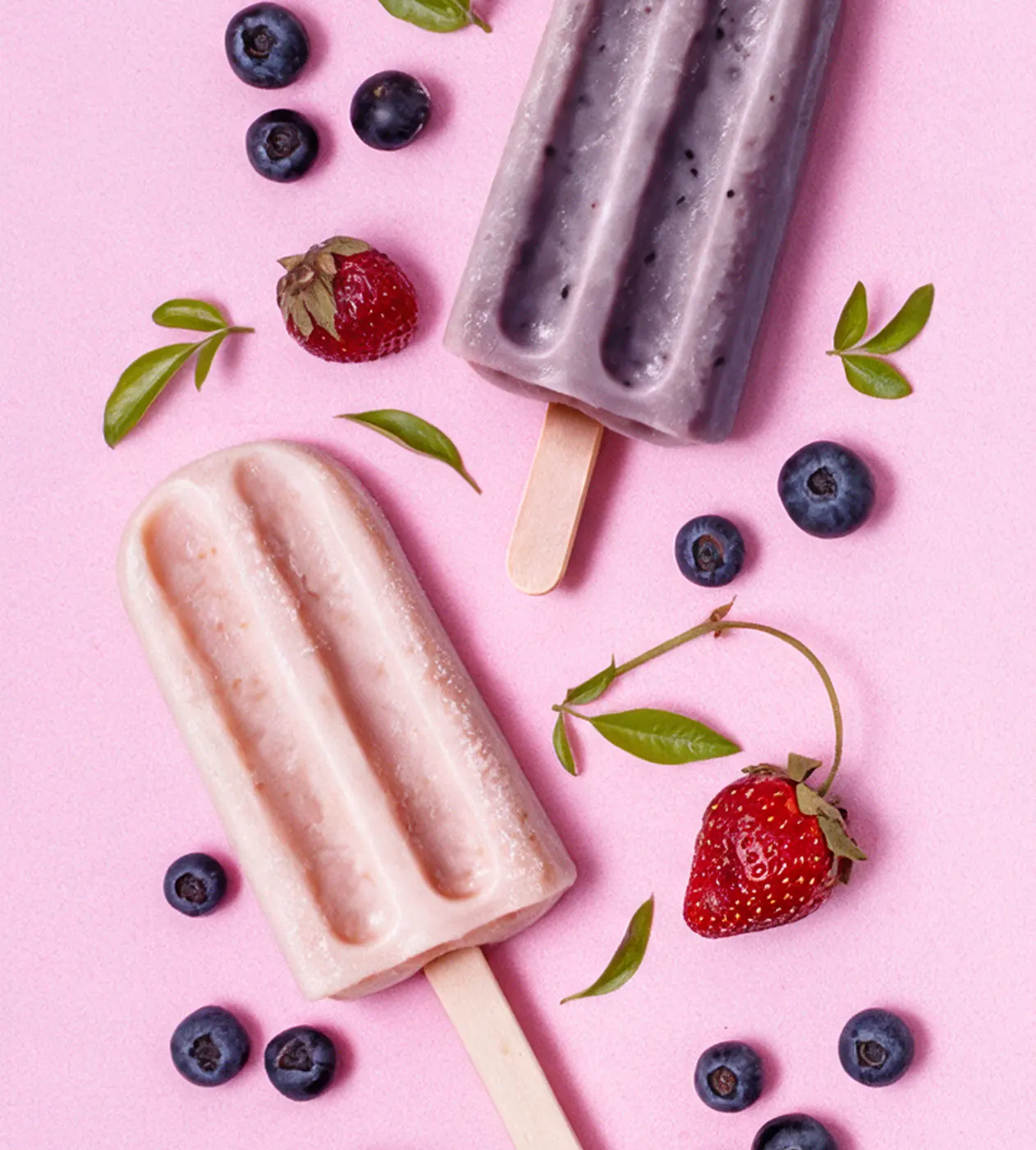 A strawberry and a blueberry popsicle on a pink table scattered with berries and mint leaves.